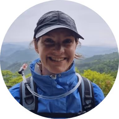 A woman in a blue jacket with a beautiful view behind her smiles at the camera